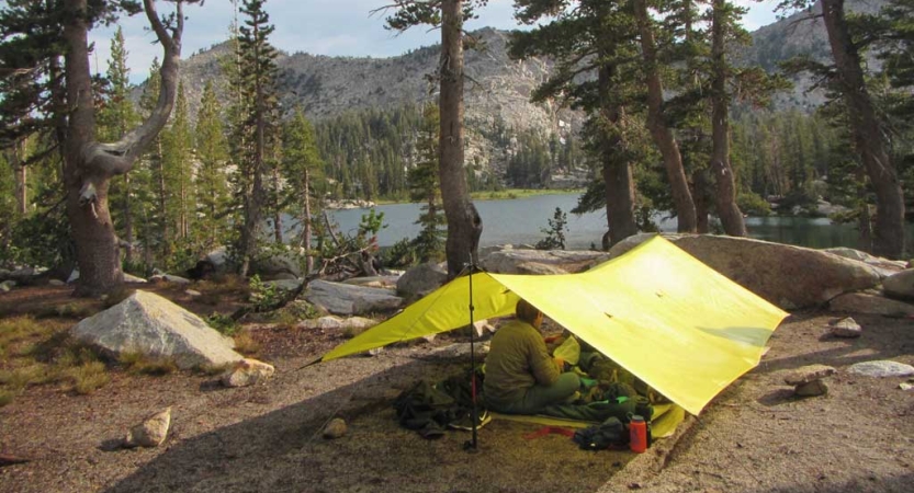 A person sits under a yellow tarp shelter that rests beside a body of water. There are mountains in the background. 